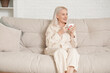 © Pixel-Shot - Mature woman in pajamas with coffee cup sitting on sofa at home