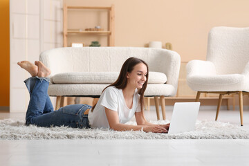 Wall Mural - Young happy woman lying on floor and using laptop near sofa in living room
