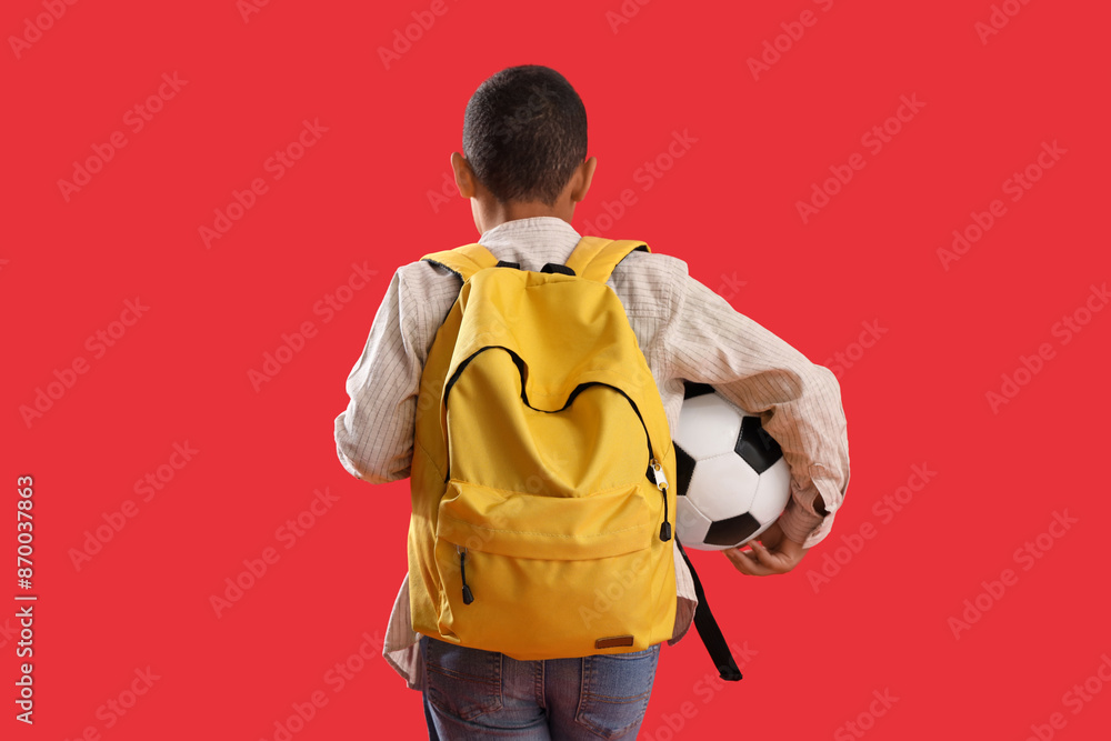Little schoolboy with backpack and soccer ball on red background, back view