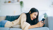 © Monster Ztudio - Happy young asian woman relax on comfortable couch at home texting messaging on smartphone, smiling girl use cellphone, browse wireless internet on gadget, shopping online from home