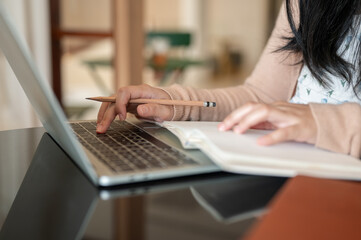 A cropped image of a woman working on her laptop, taking notes in a book and typing on the keyboard.