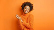 © Aytana - Pretty curly haired young woman smiles gently keeps hands together focused directly at camera has pleasant talk with best friend dressed in casual sweater isolated over vivid orange background