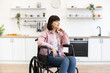 © sofiko14 - Woman sitting in wheelchair in modern kitchen setting with shelves and plants. She looks thoughtful and at ease, conveying independence and comfort.
