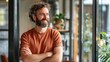 © liliyabatyrova - A man with curly hair and a beard stands in a cafe, smiling confidently and looking to the side. He is leaning against a window, arms crossed, in a casual orange shirt