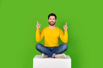 Wall Mural - Full length photo of handsome young guy point up empty space sit cube dressed stylish yellow garment isolated on green color background