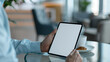 © Jane - man in a light blue button-up shirt, sitting at a glass-top table in a cafe with a coffee cup and pastry, holding a black tablet iPad Pro vertical with white blank screen, mockup