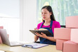 © NanSan - A woman in overalls sells products online, inspects parcels, takes customer orders, works at a desk, and uses pink boxes to pack items ready for shipping to customers.