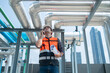 © Washburn - A construction engineer in a safety vest and helmet inspects industrial pipes on a rooftop while communicating via walkie-talkie.