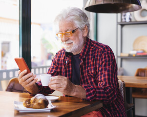 Sticker - Smiling senior bearded man sitting at cafe table using mobile phone while enjoying break with espresso coffee cup and croissant. Breakfast time