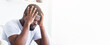 © Prostock-studio - A young black man with short, dark hair is sitting indoors, wearing a white t-shirt. He is holding his head in his hands, looking down. He appears to be distressed or overwhelmed, copy space