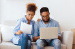 © Prostock-studio - Buying House. Serious Black Couple Using Laptop Reading Property Ownership Papers Sitting On Sofa In New Home. Empty Space