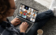 © Prostock-studio - A man sits on the floor with his legs crossed, using a laptop to participate in a group video call. The laptop screen shows a grid of multiple participants in the call.