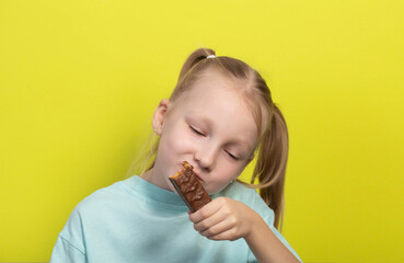 A beautiful joyful girl with white hair of seven years old eats a chocolate bar on a yellow background. Children are lovers of sweet junk food.