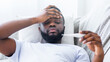 © Prostock-studio - African American man with a beard lies in bed with a blanket covering him. He holds a thermometer in his hand and has his other hand on his forehead, looking down and appearing unwell.