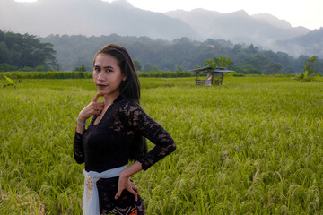 a beautiful Indonesian woman wearing a black Balinese kebaya with a background of rice fields and mountains