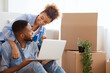 © Prostock-studio - Excited Afro Spouses Using Laptop Computer Sitting On Sofa Among Unpacked Moving Boxes In New House. Free Space, Selective Focus