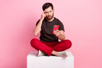 Wall Mural - Full size photo of smart man dressed brown t-shirt sit on cube look at smartphone finger on forehead isolated on pink color background