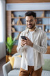 © Tetiana - Vertical shot of a young Indian man at home, standing in a room and smiling while using a mobile phone