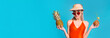 © Prostock-studio - A young woman is wearing an orange swimsuit and a straw hat, and holding a pineapple in one hand and a coconut with a straw in the other. She is smiling and looking directly at the camera.