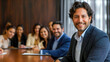 © Ron - smiling business man in his late thirties sitting at a table with a group of diverse people, in a modern board room background