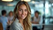 © Pinklife - A woman with blonde hair and blue eyes smiling confidently while sitting at an office desk, with blurred colleagues in the background enhancing depth and context.