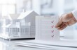 © KanitChurem - A hand holding a checklist with red checkmarks, in front of a house model on a desk.