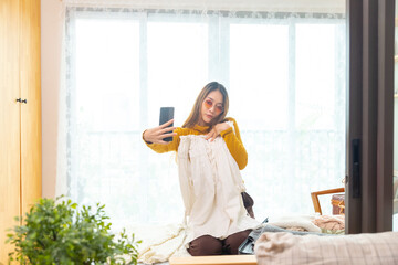 Happy Asian woman using smartphone taking selfie during packing clothes in suitcase in bedroom. Attractive girl traveler enjoy outdoor lifestyle preparing for travel trip on summer holiday vacation.