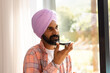 © Wavebreak Media - Speaking into smartphone, man in turban standing by window at home