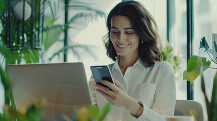 Sticker - A woman in a white blouse smiles as she checks her phone, sitting at a desk with a laptop amidst lush green plants in a bright, modern workspace.