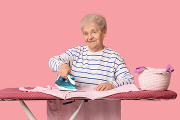Wall Mural - Happy senior woman ironing laundry on pink background