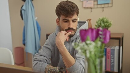 Poster - A thoughtful hispanic man with a beard contemplates indoors, surrounded by books, flower decor, and casual home attire.