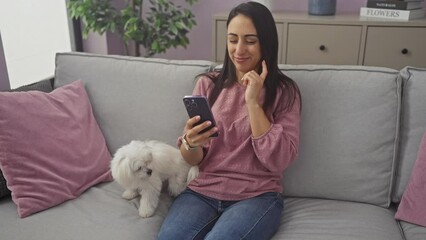 Wall Mural - A young hispanic woman in casual clothing uses a smartphone while sitting on a couch next to a white bichon maltese dog inside a cozy living room