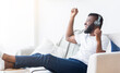© Prostock-studio - African American man wearing headphones is sitting on a couch and listening to music. He is smiling and has his hand raised in the air, as if he is dancing or singing along to the music.