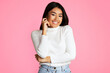 © Prostock-studio - Asian young woman with long black hair is smiling brightly, wearing a white sweater and blue jeans. She is standing against a solid pink background. Her right hand is resting on her cheek