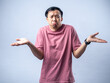 © Daniel - An Asian man in a pink shirt shrugs with a confused and uncertain expression, his arms outstretched and palms up. The plain light blue background emphasizes his ambiguous