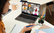 © Prostock-studio - This image shows a woman sitting at a desk, participating in a video conference meeting on her laptop. The screen displays multiple participants in a grid format, suggesting a virtual team meeting or