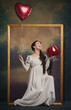© master1305 - Happy smiling woman in period dress sitting in picture frame with heart-shaped balloon and wine glass on vintage background. Birthday celebration. Concept of history, comparison of eras, remake