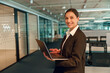 © Viktoria Kovalchuk - Confident businesswoman is smiling and holding a laptop in a modern open office workspace