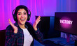 © Prostock-studio - A young woman wearing headphones sits at a computer desk in a dimly lit room, illuminated by pink and blue lights. She is smiling and raising her hands in the air, celebrating her victory.