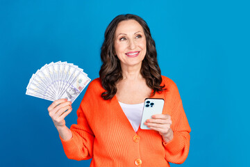 Poster - Photo of attractive senior woman hold device money fan look empty space dressed stylish orange clothes isolated on blue color background