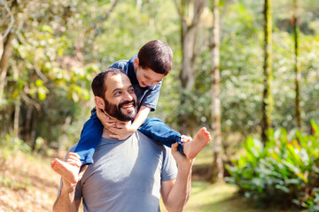 A joyful Brazilian father carrying his young son on his back in a lush, green forest. They are laughing and playing, highlighting a special moment of father-son bonding on Father's Day	