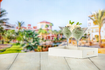 Poster - Two glass cups of vanilla and mint ice cream on wooden board table on sandy tropical beach 