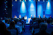 © sanjit536 - Stylish business event, panel discussion on stage with audience in foreground, dark and blue and dark blue background