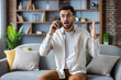 © Tetiana - Portrait of shocked and worried young Indian man sitting on sofa at home, talking on mobile phone, throwing up hands in frustration