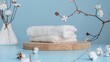 © liliyabatyrova - Two white, fluffy towels are stacked on top of a wooden round cutting board. The background is a light blue. There are several cotton flower stems on the table