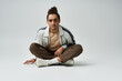 © LIGHTFIELD STUDIOS - A young African American man sits cross-legged on a gray background wearing a stylish white jacket, brown pants, and white sneakers.
