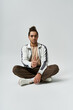© LIGHTFIELD STUDIOS - A young African American man sits with his legs crossed, wearing a white jacket and brown pants, against a gray background.