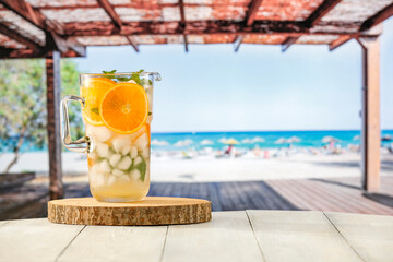 Sticker - Glass of mocktail drink with ice and citrus fruit on a wooden table with sandy tropical beach and blue sky background. Summer table and copy space for products and food and drinks and fruit.