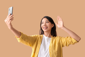 Poster - Beautiful young Asian woman with mobile phone waving hand on beige background