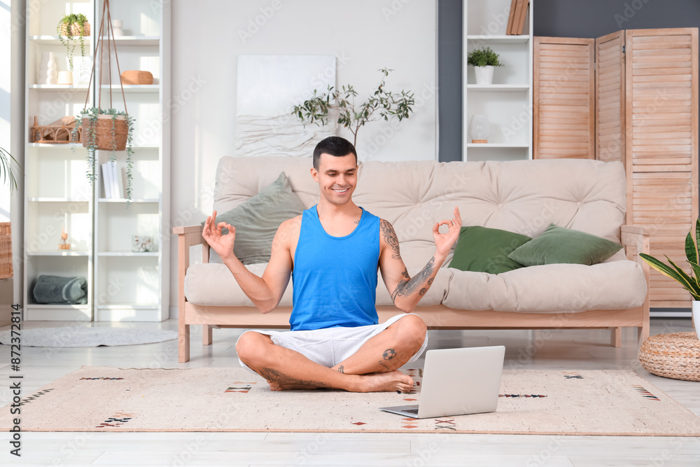 Young man with laptop doing yoga on carpet at home